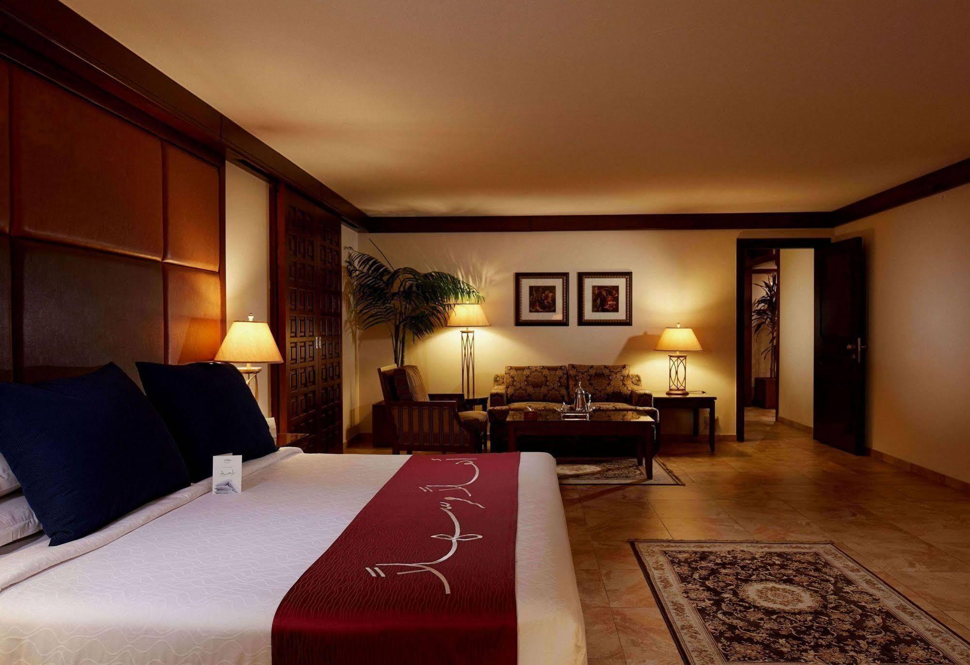 Makarem Ajyad Makkah Hotel Mecca Екстер'єр фото The photo depicts a cozy hotel room with a warm and inviting atmosphere. It features a large bed with dark blue pillows and a maroon throw blanket. On either side of the bed, there are lamps providing soft lighting. In the background, a small seating