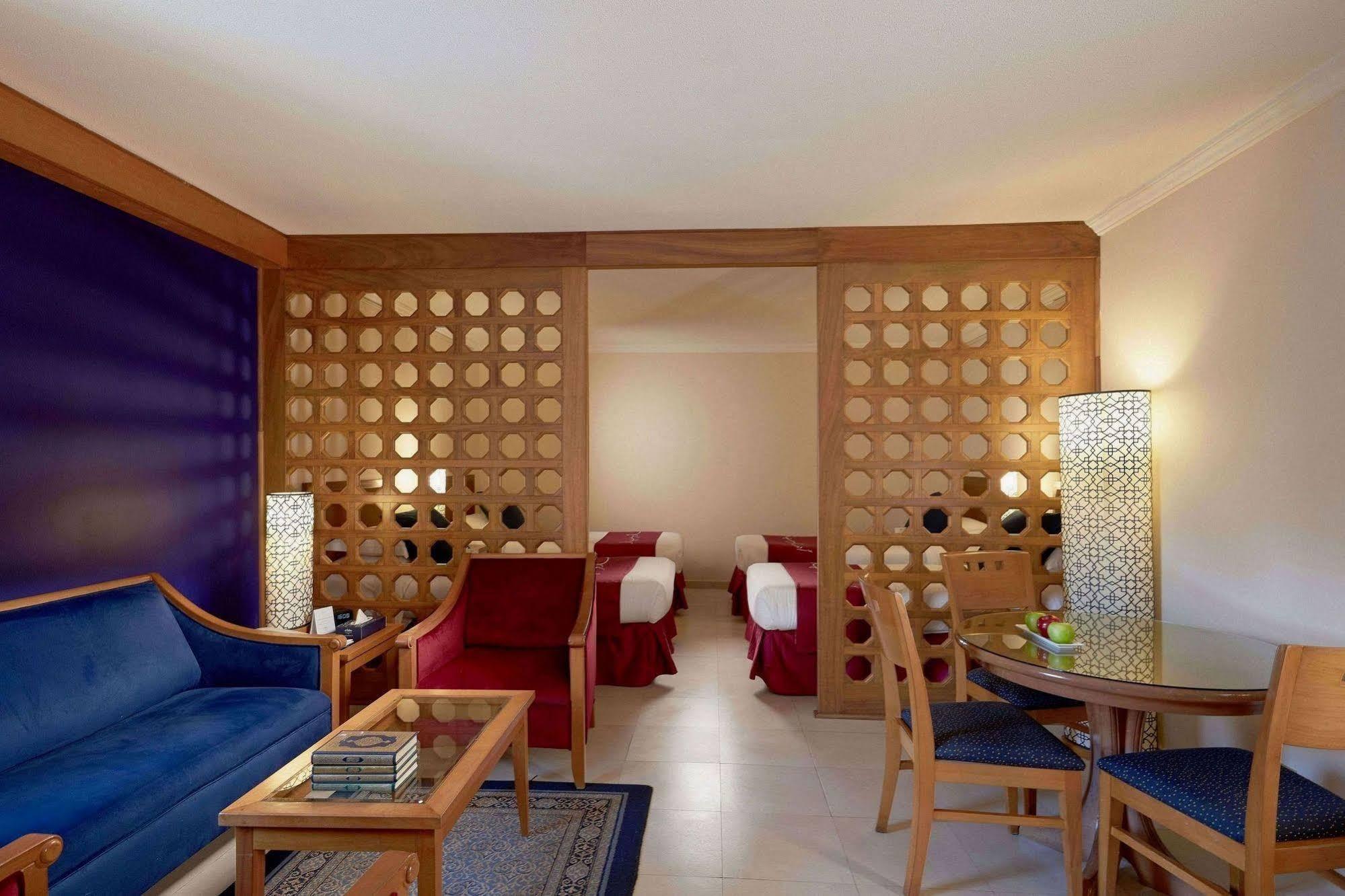 Makarem Ajyad Makkah Hotel Mecca Екстер'єр фото The photo shows a well-furnished hotel room or suite. On the left side, there is a seating area featuring a blue sofa and a small coffee table, as well as a couple of decorative items. The room is separated by a decorative wooden partition with circu
