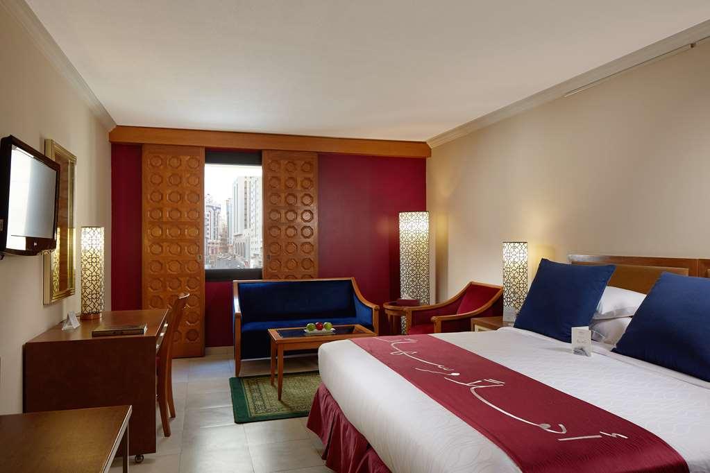 Makarem Ajyad Makkah Hotel Mecca Номер фото The photo showcases a hotel room featuring a modern design. In the center, there's a large bed with white linens and a decorative red runner. On either side of the bed, there are wooden nightstands with lamps. A blue sitting area is visible, composed