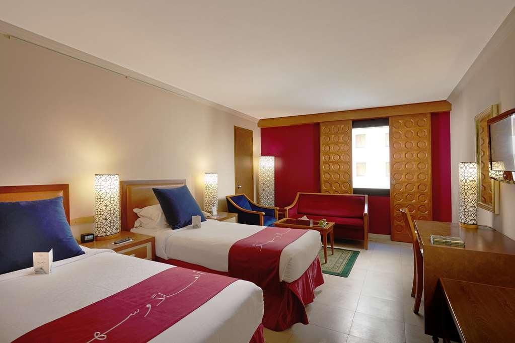 Makarem Ajyad Makkah Hotel Mecca Номер фото The photo shows a hotel room featuring two twin beds with red and white bedding. There are decorative lamps on either side of the beds. A small seating area is visible, including a blue chair and a red sofa. There is a wooden desk against one wall, a