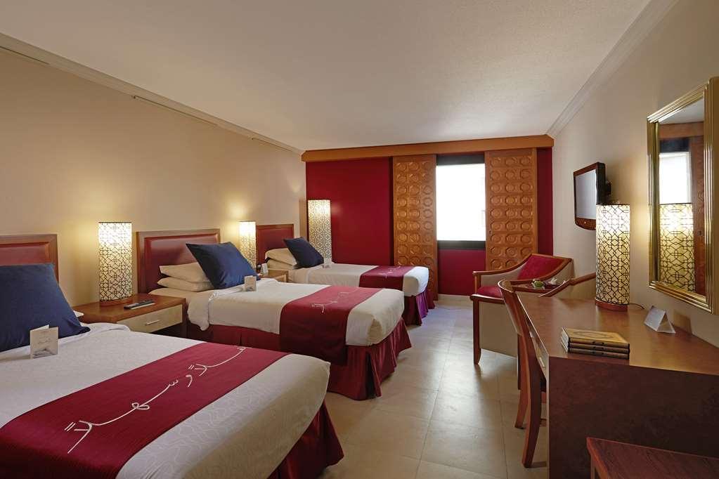 Makarem Ajyad Makkah Hotel Mecca Номер фото The photo shows a hotel room featuring three beds arranged with red and white bedding. The walls are painted in a neutral tone, and there are decorative lamps on nightstands beside each bed, providing ambient lighting. One of the walls has a window c