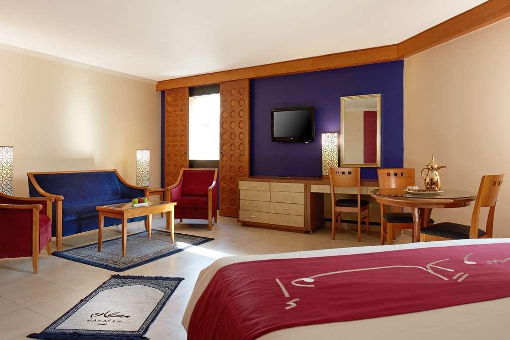 Makarem Ajyad Makkah Hotel Mecca Номер фото The image shows a hotel room with a modern, comfortable design. The room features a large bed with a red and white bedspread, a seating area that includes a blue sofa, two red chairs, and a wooden coffee table. There's a flat-screen TV mounted on the