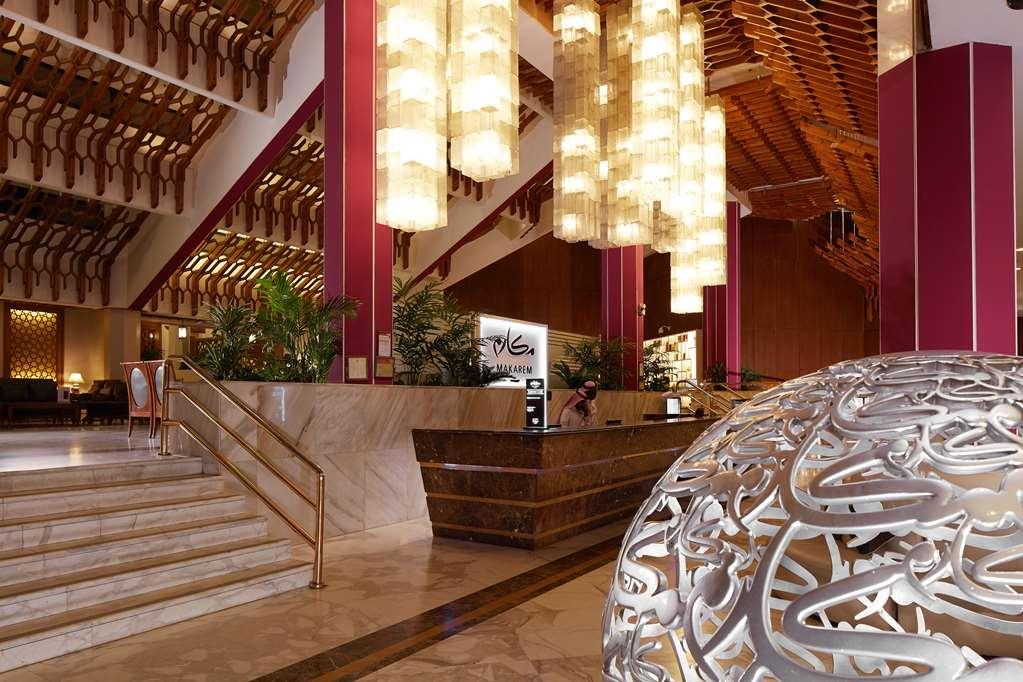 Makarem Ajyad Makkah Hotel Mecca Інтер'єр фото The photo depicts a luxurious hotel lobby. It features a grand entrance with a staircase leading up to a reception area. The lobby has high ceilings adorned with striking, modern light fixtures that hang down, providing a warm ambiance. The walls are