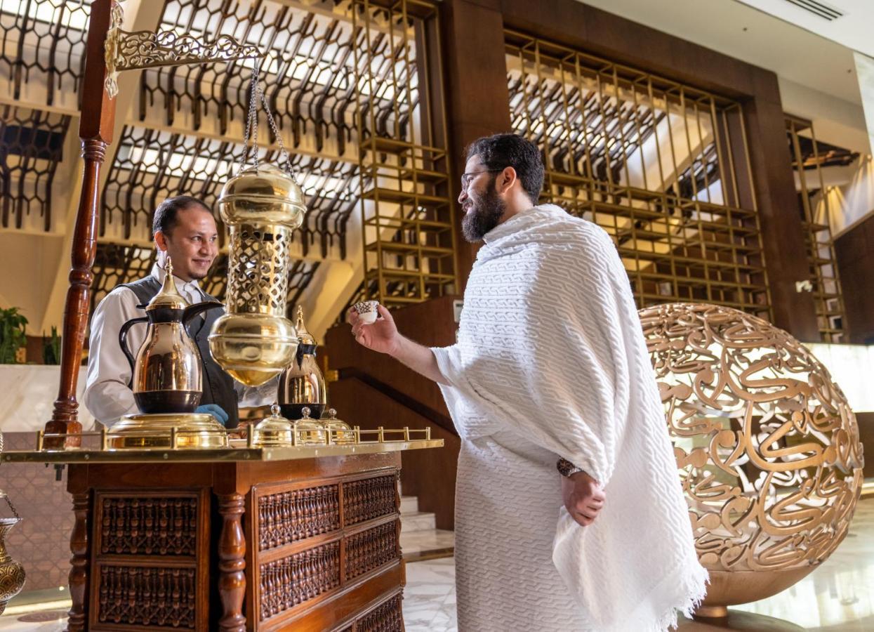 Makarem Ajyad Makkah Hotel Mecca Екстер'єр фото The photo depicts a scene in a luxurious setting, likely a hotel or a fine dining establishment. In the foreground, a man wearing traditional attire, possibly a white garment, is engaging with a staff member. The staff member is behind a wooden count