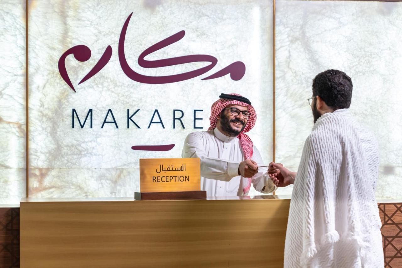 Makarem Ajyad Makkah Hotel Mecca Екстер'єр фото The photo shows a reception area with a sign that reads "MAKARE" in both Arabic and English. In the foreground, a staff member wearing traditional attire is smiling and shaking hands with a visitor, who is clad in a white garment, typical for formal 