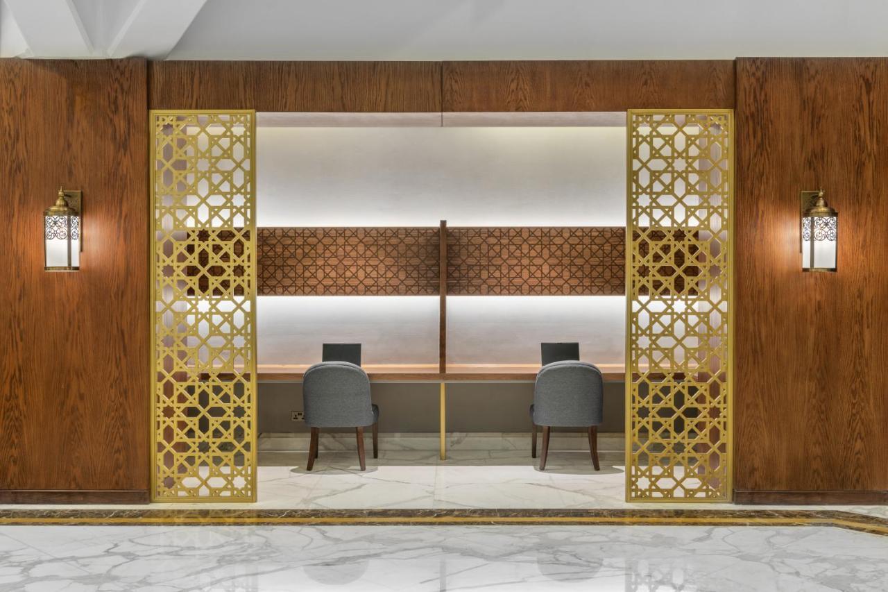 Makarem Ajyad Makkah Hotel Mecca Екстер'єр фото The photo shows an elegantly designed space that features a reception or working area. There are two workstations with chairs positioned in front of them. The backdrop has a decorative panel with a geometric pattern. The area is framed by ornate gold