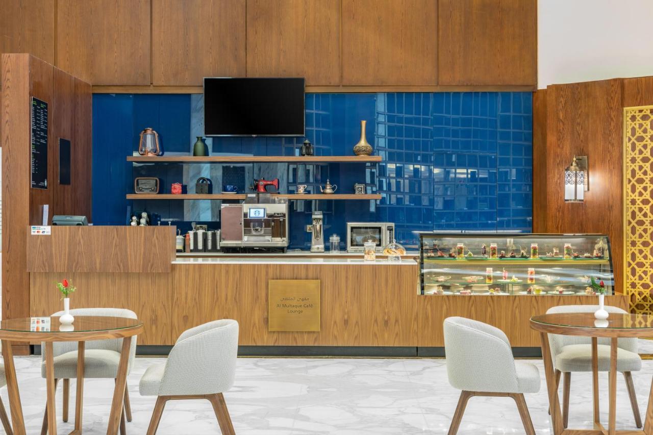 Makarem Ajyad Makkah Hotel Mecca Екстер'єр фото The photo shows the interior of a café. The design features warm wood paneling and a striking blue tiled wall. In the foreground, there are several light-colored chairs and small tables arranged neatly. Behind the counter, there's a variety of coffee