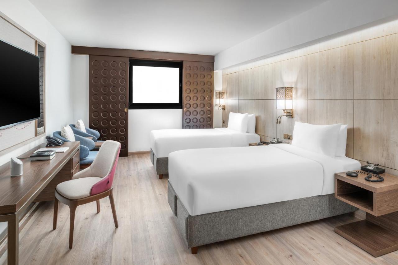 Makarem Ajyad Makkah Hotel Mecca Екстер'єр фото The photo shows a modern hotel room featuring two beds with white linens. There is a wooden accent wall with a window above it. Opposite the beds, a wall-mounted television is visible. A stylish wooden desk with a chair is situated on one side of the