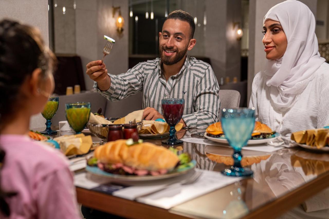 Makarem Ajyad Makkah Hotel Mecca Екстер'єр фото The photo shows a dining scene with three people enjoying a meal together. A man is seated at the table, smiling and holding a utensil, possibly ready to serve or share food. He is dressed in a striped shirt. Across from him is a woman wearing a whit