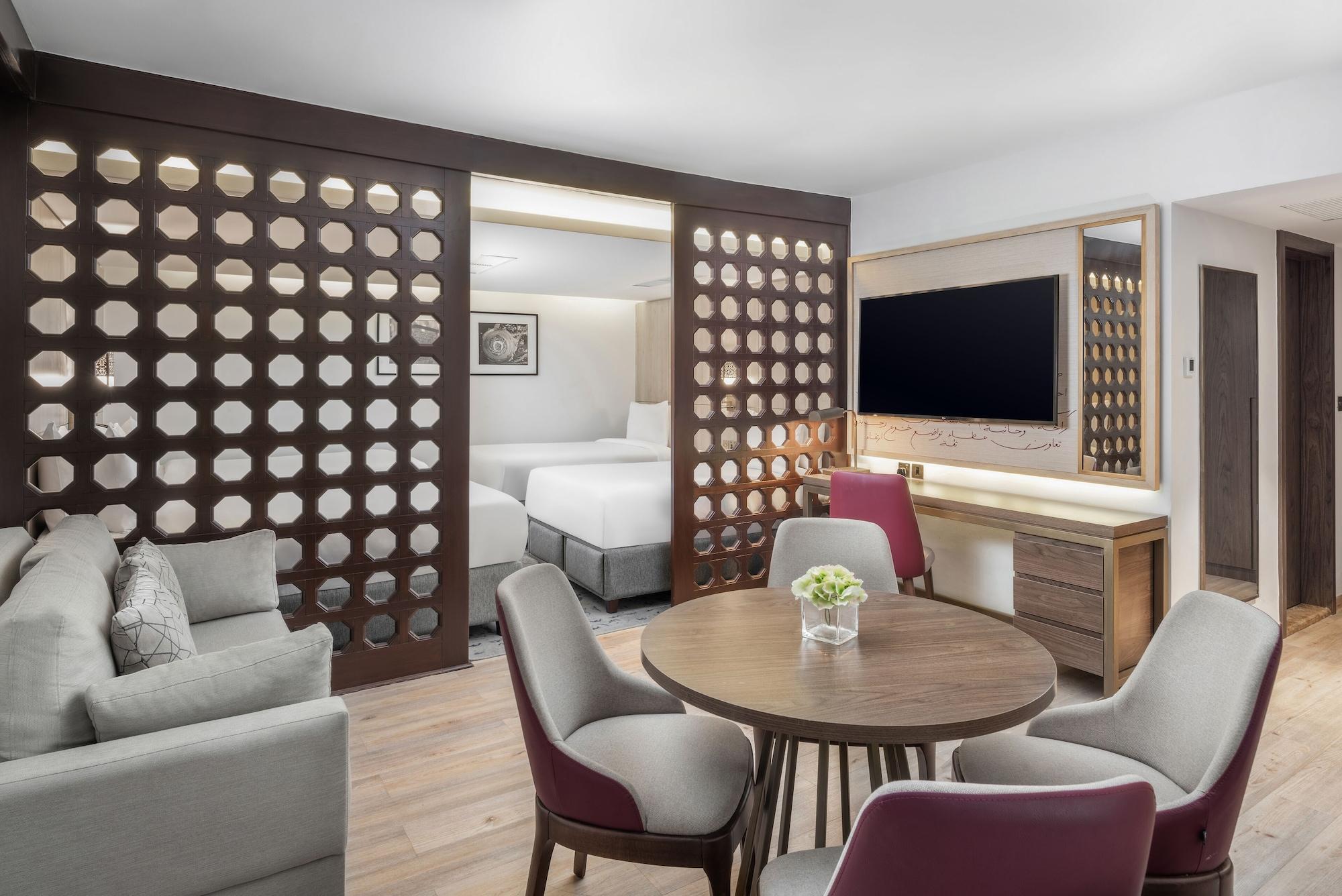 Makarem Ajyad Makkah Hotel Mecca Екстер'єр фото The photo shows a modern hotel room or suite. It features a living area with a comfortable gray sofa and a round wooden dining table surrounded by upholstered chairs. A large flat-screen TV is mounted on the wall opposite the seating area. There's a 