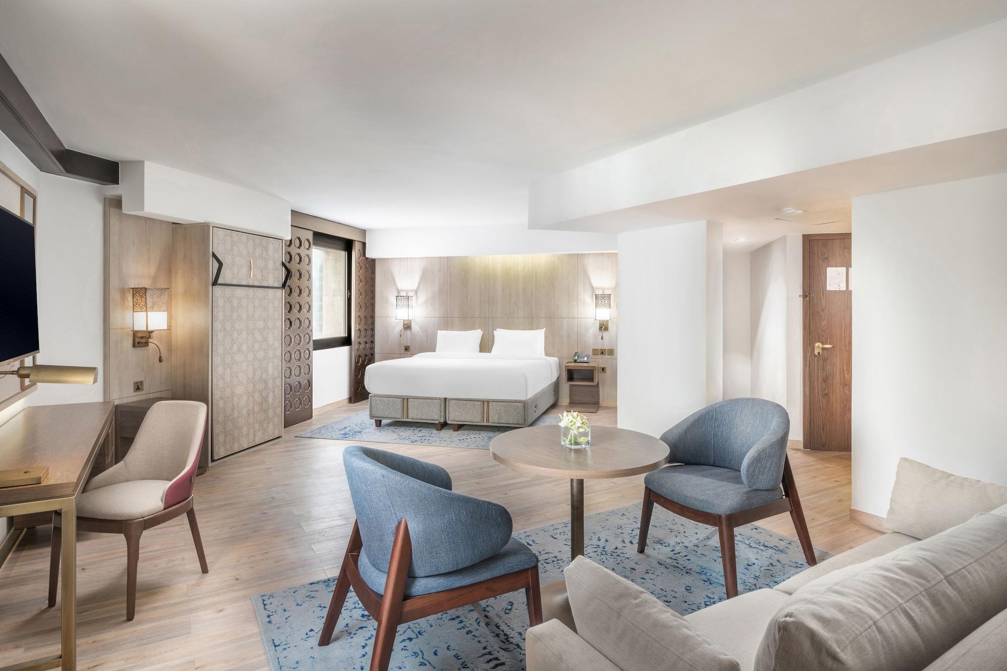 Makarem Ajyad Makkah Hotel Mecca Екстер'єр фото The photo shows a modern hotel room with a spacious layout. It features a large bed with a simple headboard against a light-colored wall. There are two soft chairs in light blue and beige positioned around a small round table. The flooring is wooden,