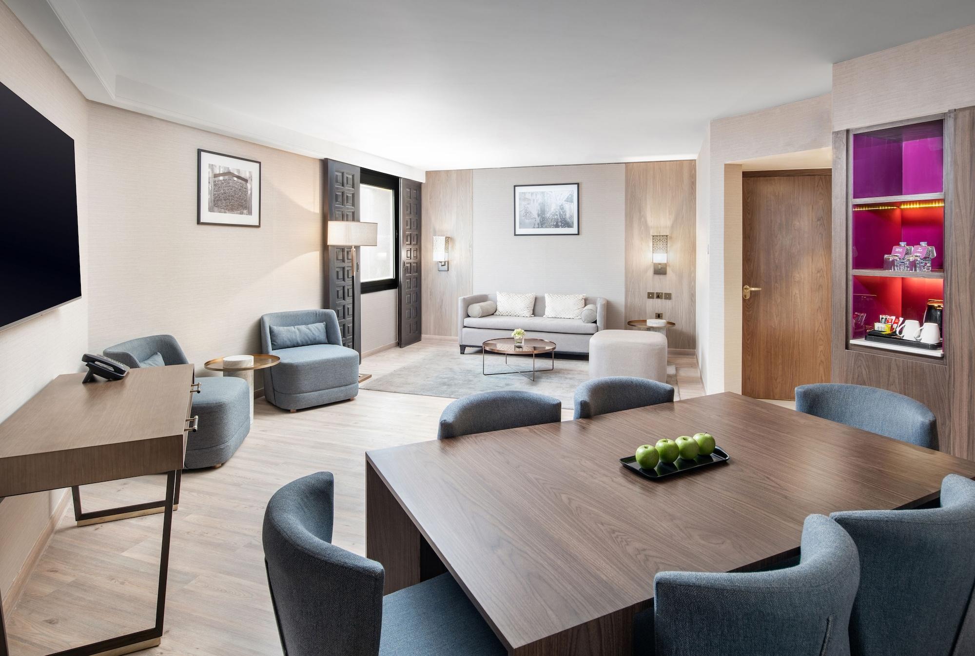 Makarem Ajyad Makkah Hotel Mecca Екстер'єр фото The photo shows a modern and elegantly furnished living area. It features a spacious layout with a comfortable seating arrangement that includes a sofa and armchairs. There is a large wooden dining table with several chairs around it. The walls are a