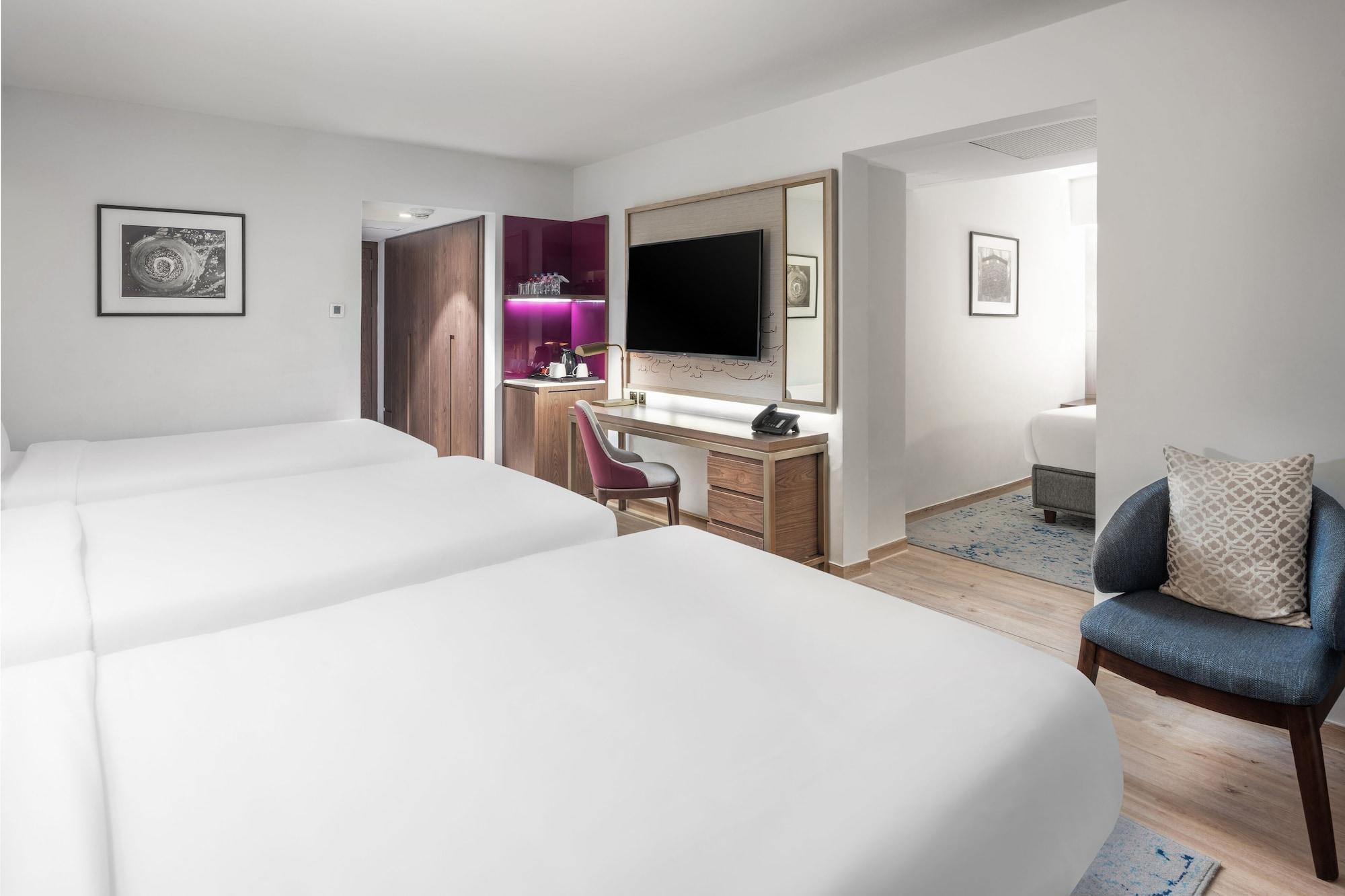 Makarem Ajyad Makkah Hotel Mecca Екстер'єр фото The photo shows a modern hotel room featuring two white beds with smooth linens. The room has a minimalistic design with light-colored walls and wooden flooring. On one side, there is a desk and chair next to a television mounted on the wall, along w