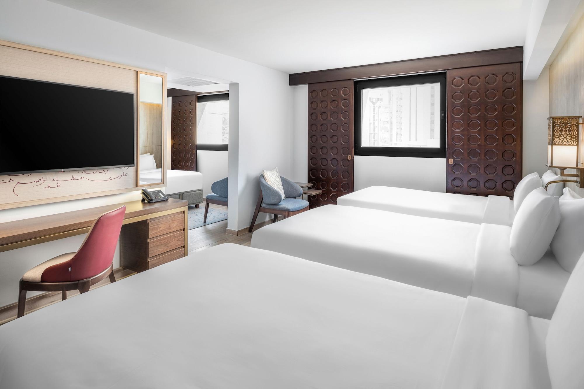 Makarem Ajyad Makkah Hotel Mecca Екстер'єр фото The photo shows a hotel room featuring three beds with white linens. There is a large flat-screen TV mounted on the wall opposite the beds, and a wooden side table beneath it. A red chair is positioned near the TV, while a blue upholstered chair is p