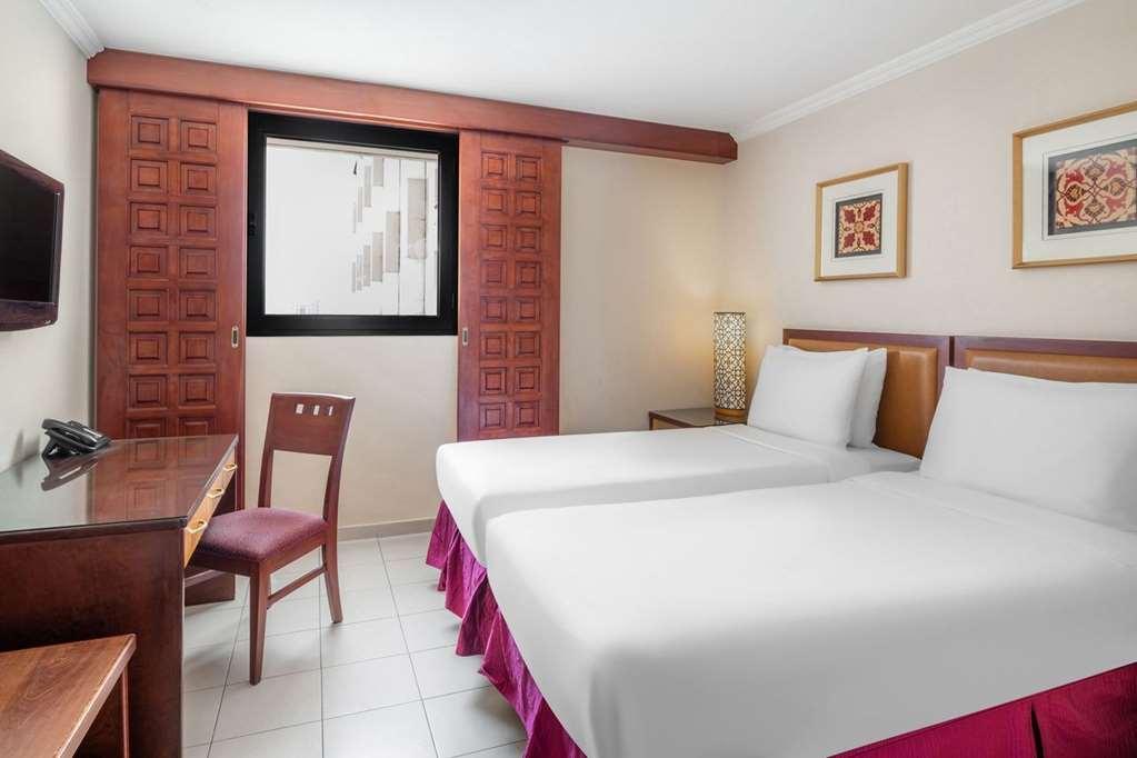 Makarem Ajyad Makkah Hotel Mecca Номер фото The photo shows a hotel room with two beds, each covered with white bedding. There is a wooden desk and a chair near the beds. The room features a window with a wooden frame and a decorative lamp beside the beds. On the wall, there are two framed art
