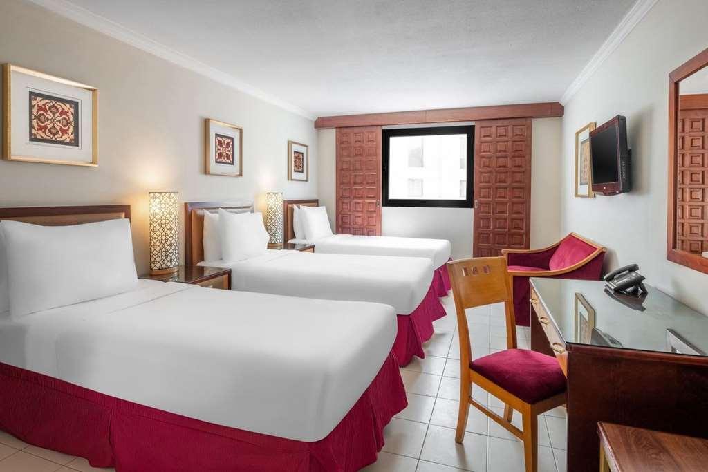Makarem Ajyad Makkah Hotel Mecca Номер фото The photo shows a hotel room featuring two twin beds with white linens and red bed skirts. There are decorative elements on the walls, including framed artwork. A large window provides natural light, while a desk and a chair are positioned next to it