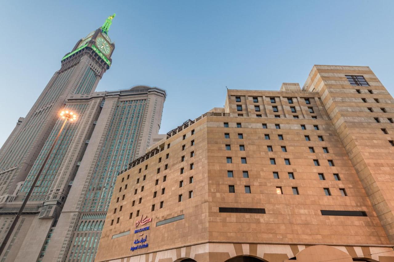 Makarem Ajyad Makkah Hotel Mecca Екстер'єр фото The photo shows a prominent building with a modern architectural design, likely a hotel or commercial establishment, at the forefront. It features a light-colored stone facade with large windows. Behind it, there is a tall skyscraper, which has a dis