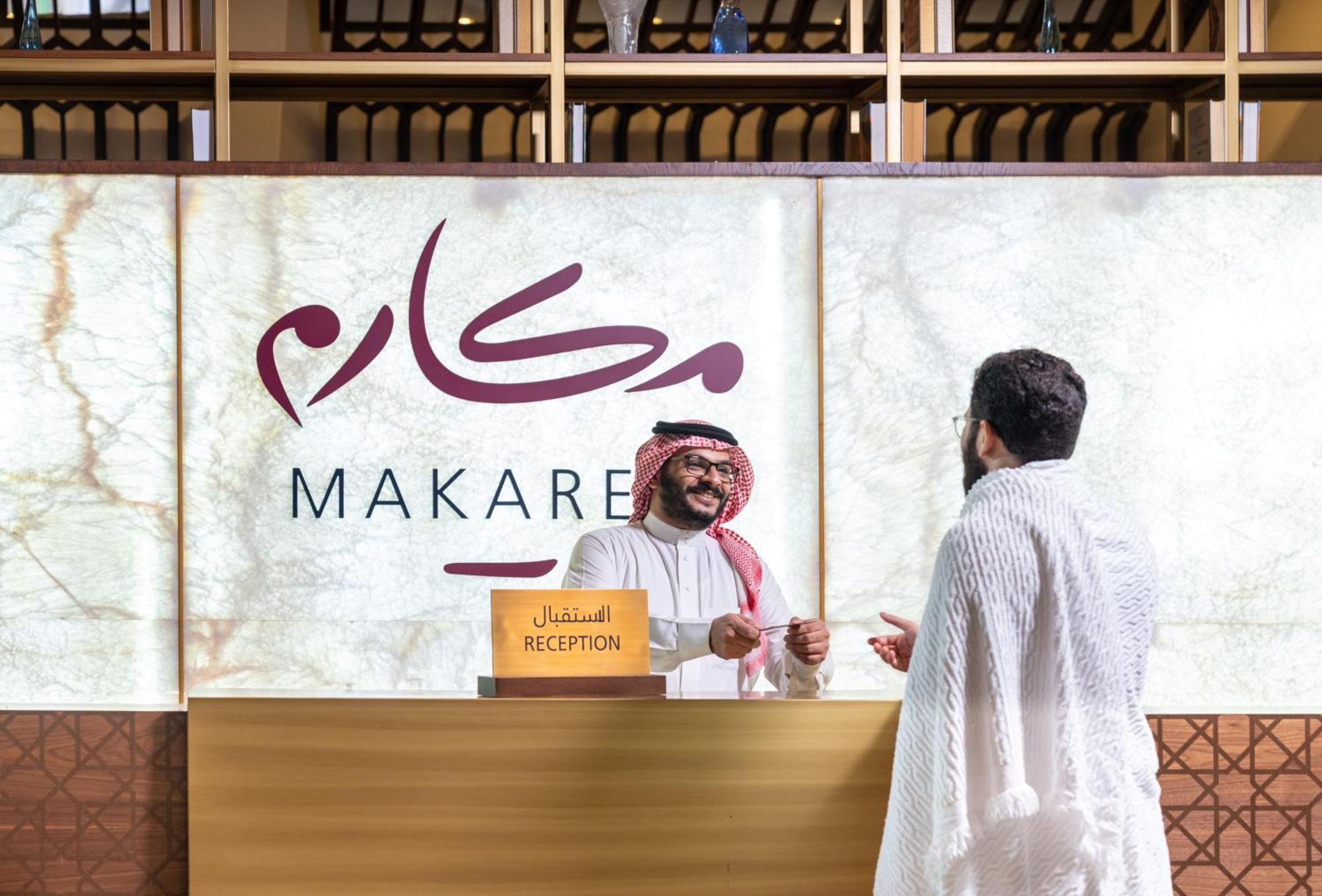 Makarem Ajyad Makkah Hotel Mecca Екстер'єр фото The photo depicts a reception area in a hotel or establishment named "Makarem." In the image, a receptionist, dressed in traditional attire, is smiling and interacting with a guest who is wearing a white robe, commonly known as a thobe or ihram. The 