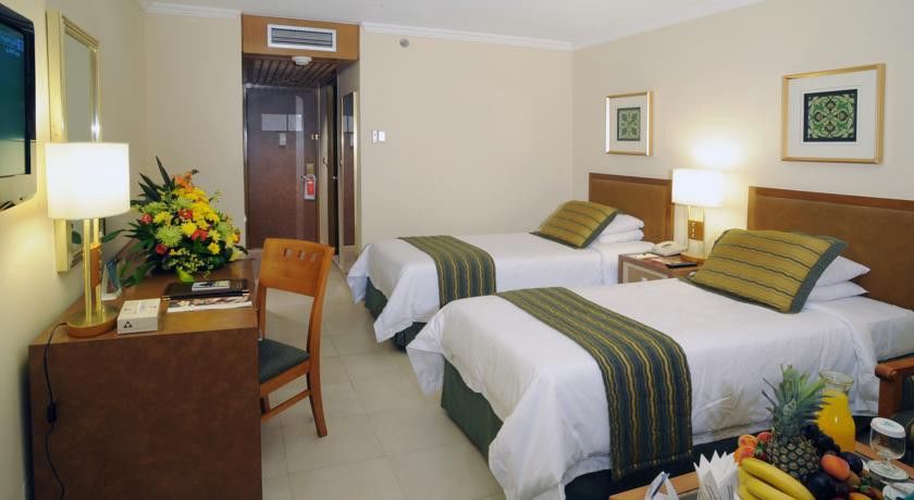 Makarem Ajyad Makkah Hotel Mecca Екстер'єр фото The photo shows an interior of a hotel room featuring two twin beds with white linens and green decorative accents. The walls are painted in a light beige color, and there is a small table next to one of the beds. A desk with a chair is positioned ne